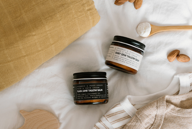 Two jars of baby balm on a white surface with wooden spoons and nuts.