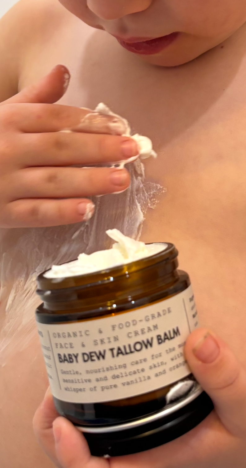 Person applying a white cream from a jar labeled 'Baby Dew Tallow Balm' to their skin.